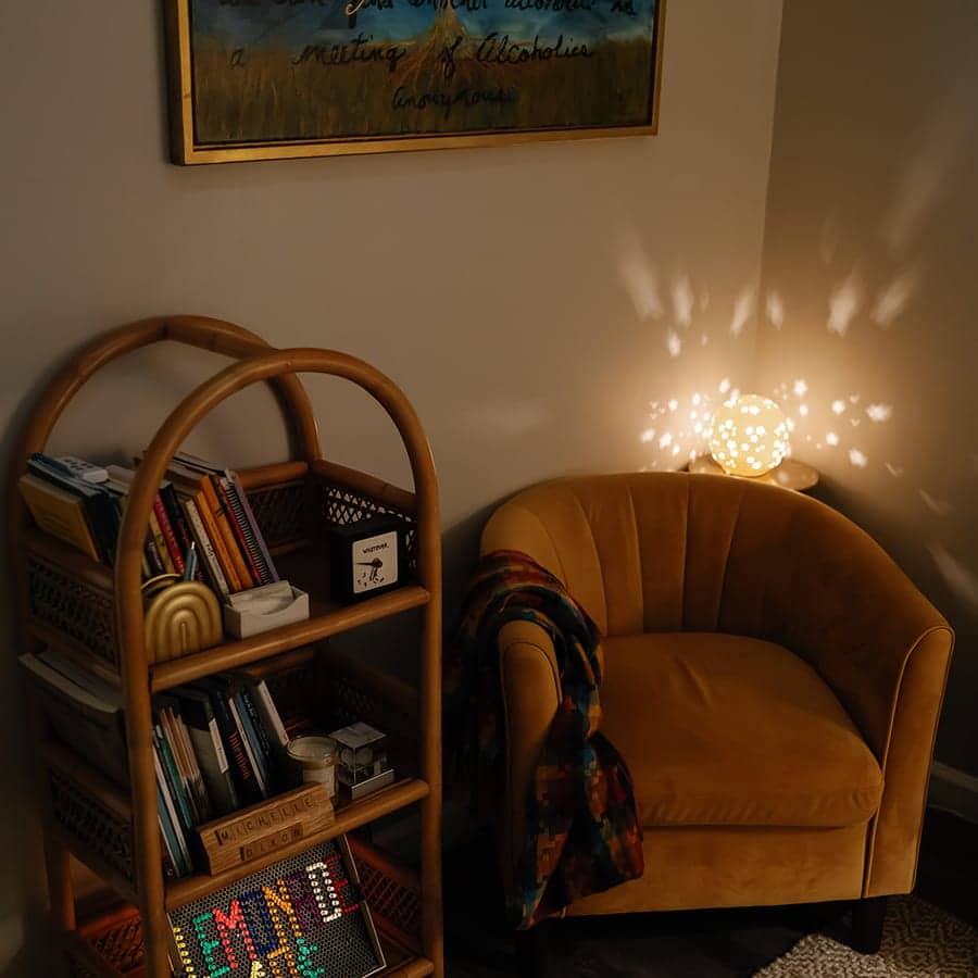 A velvet armchair beside a rattan bookshelf, lit by a lamp that scatters points of light across the wall