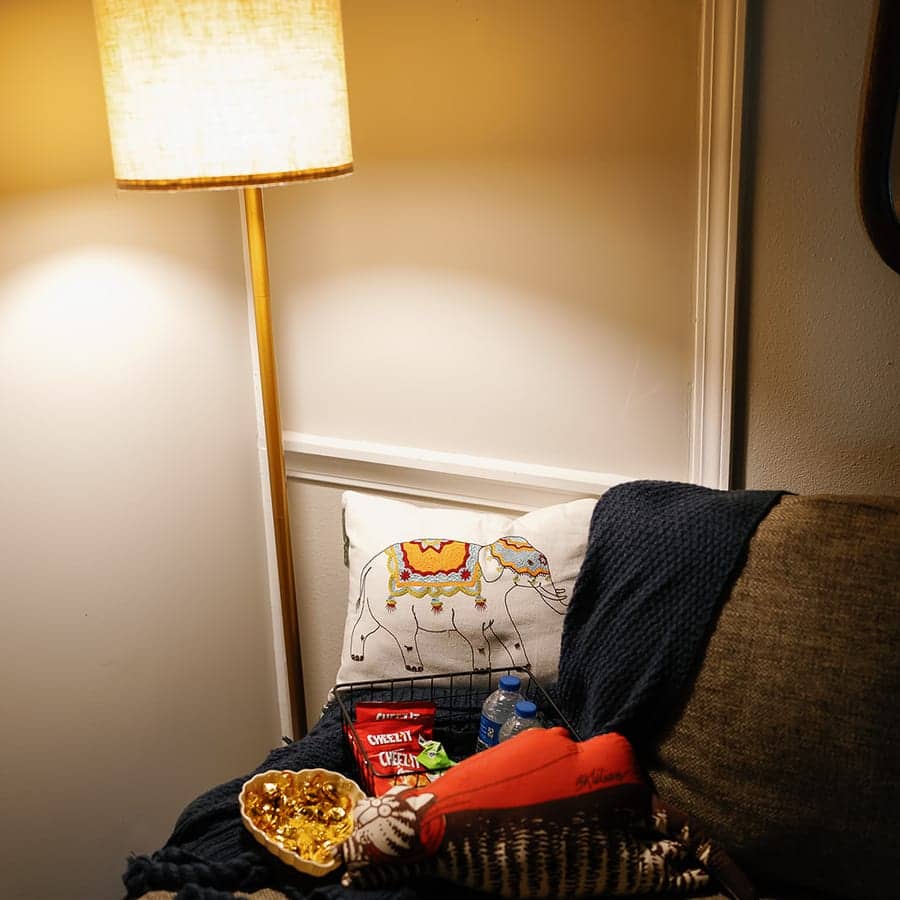 A floor lamp lit beside a couch with a folded blanket and a cushion