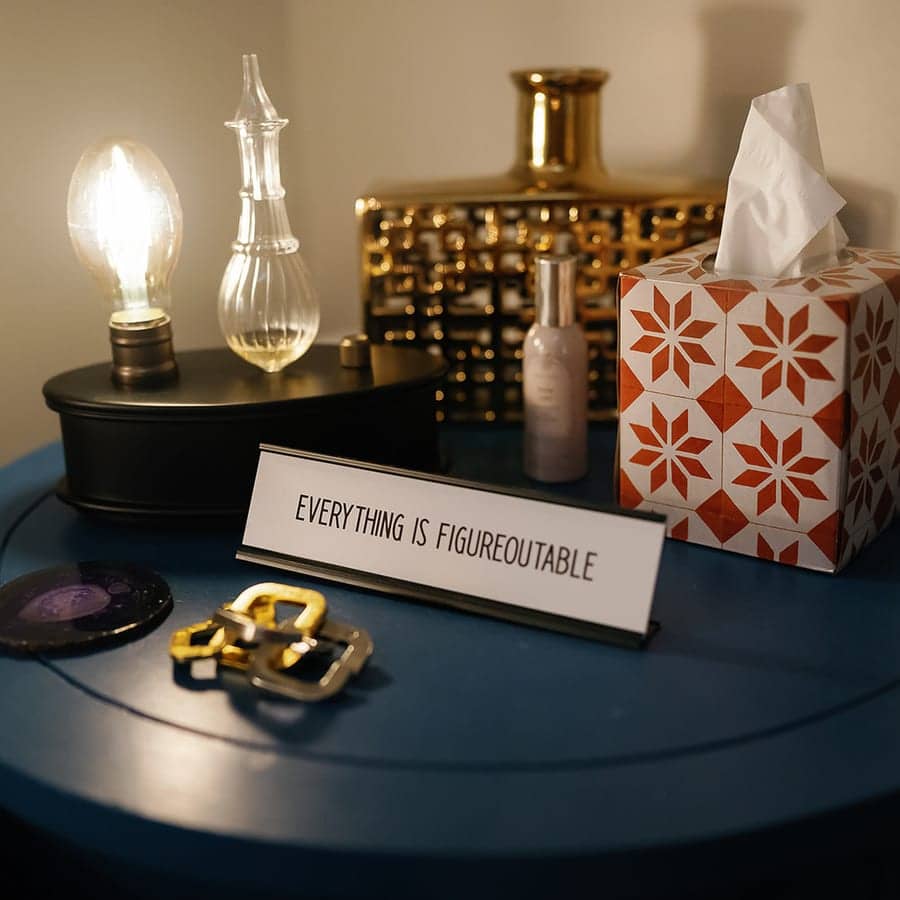 A side table with an Edison-bulb lamp, a brass vessel, a tissue box, and a small sign reading Everything Is Figureoutable